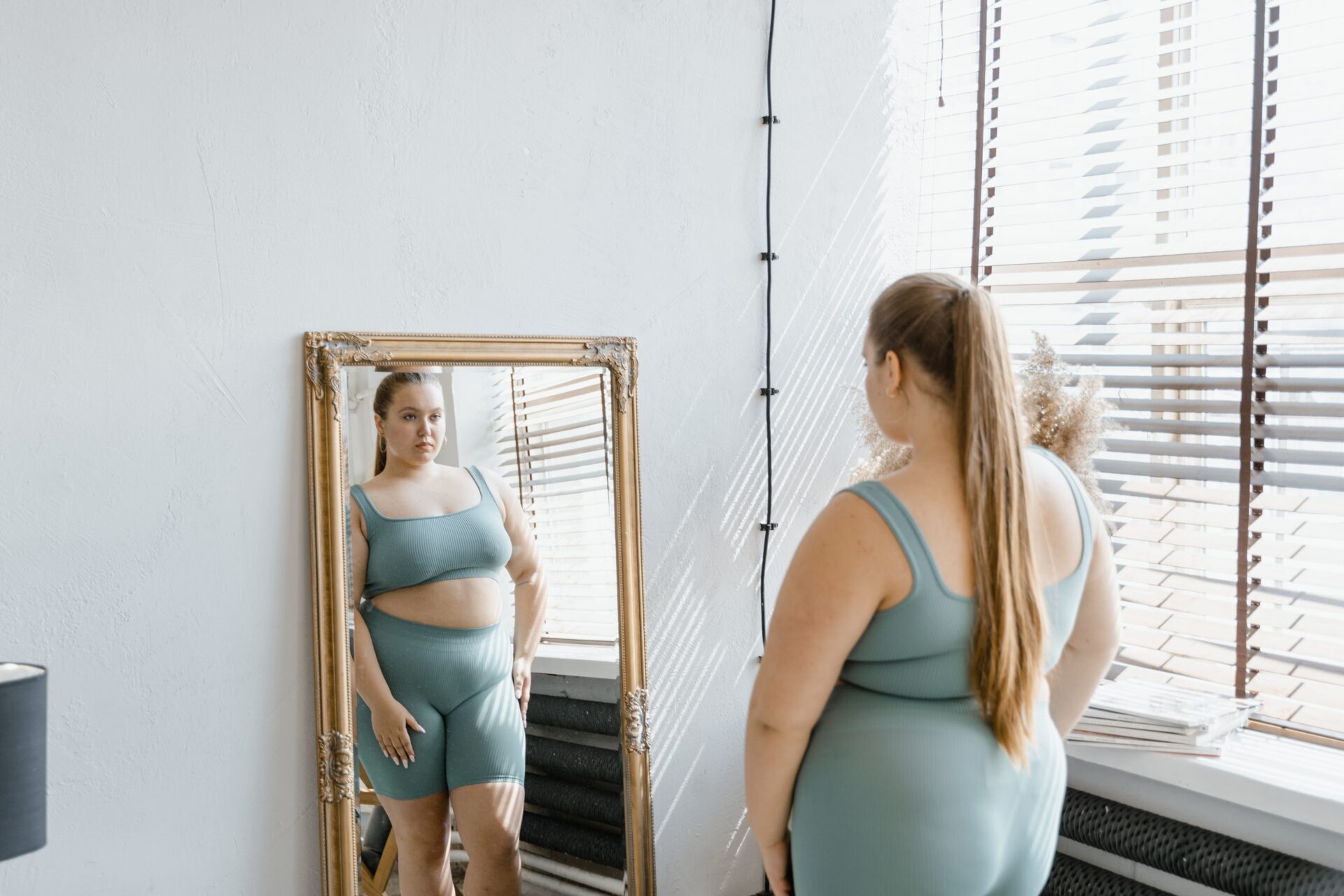 A woman in a green sports bra and shorts, at Perth Weight Surgery, gazes at herself in the mirror.